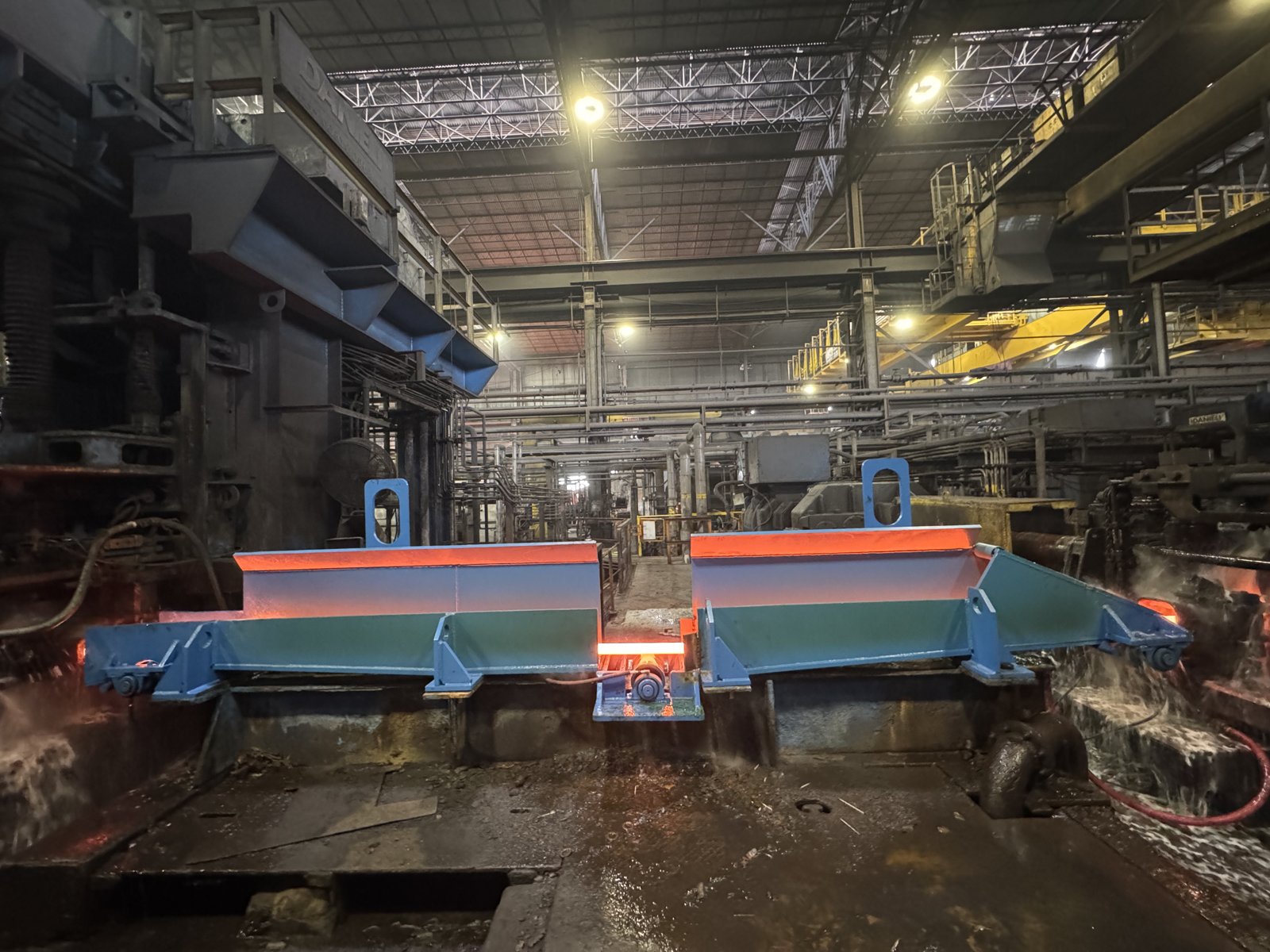 Water-cooled roller trough assembly on a hot mill line, with red-hot steel bar mid-transit through the gap between the two trough sections.