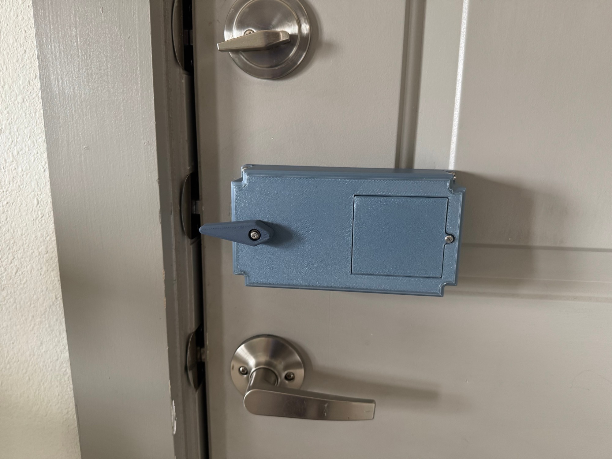 Photo of the smart door lock installed on a real door — slate-blue 3D-printed enclosure mounted between the deadbolt and the door handle, with the lever arm engaged on the latch.