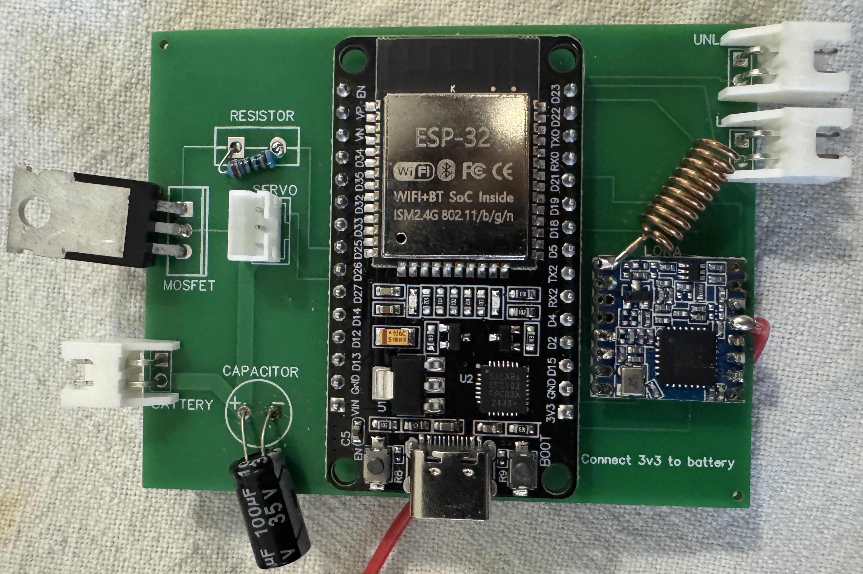 Photo of the assembled door-unit PCB — ESP32-DevKit seated in the center via header sockets, SX1276 LoRa module with helical antenna on the right, MOSFET, resistor and 100 µF bulk capacitor through-hole on the left, and JST connectors silkscreened BATTERY, SERVO, LOCK, and UNLOCK.
